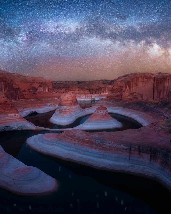 Starry nightscape over striped sandstone formations and serene water reflections.