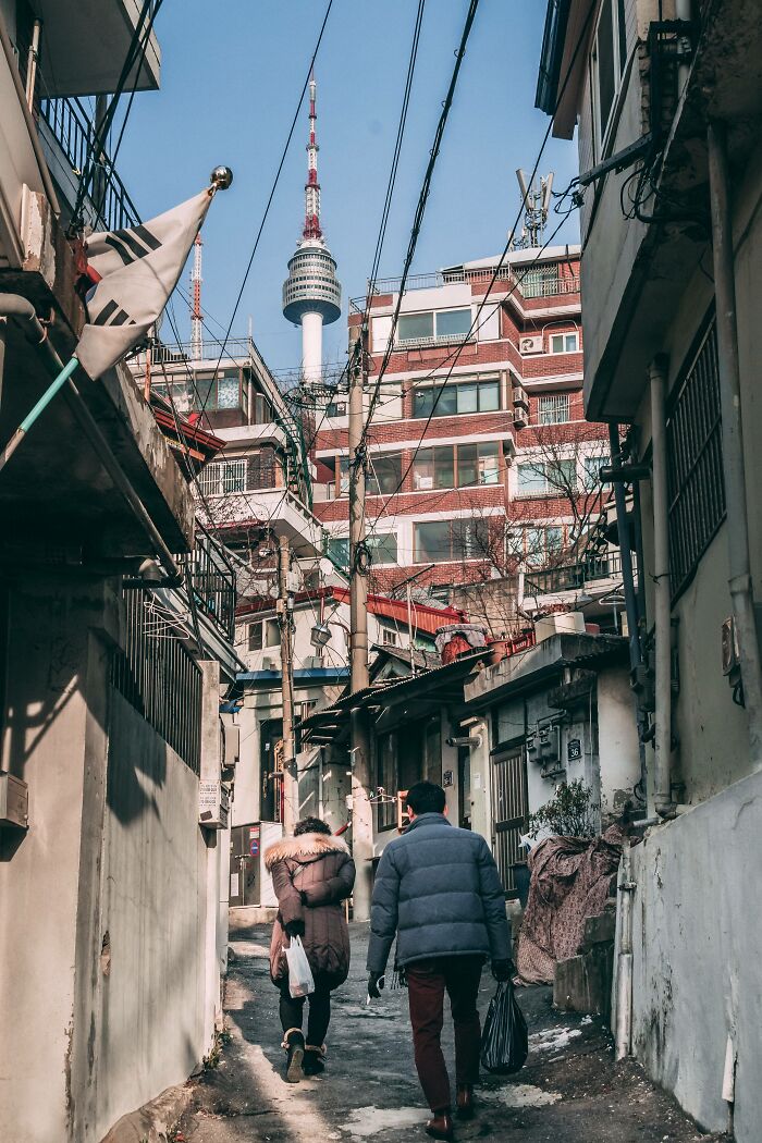 Narrow Seoul street with two people walking, traditional and modern buildings, and Namsan Seoul Tower in the background.