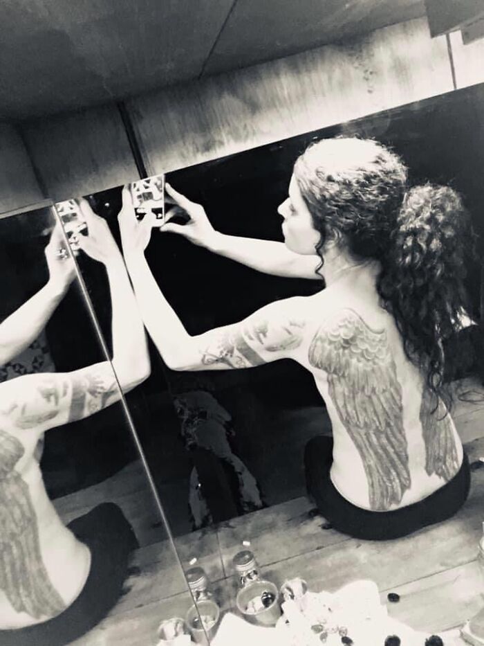 Person with wing tattoos on their back, taking a mirror selfie in a room.