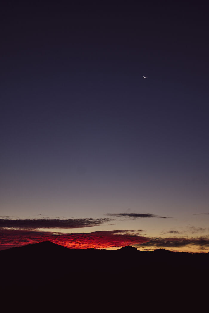 Silhouette of Ukrainian Carpathians at sunset with a crescent moon in a clear twilight sky above red clouds.