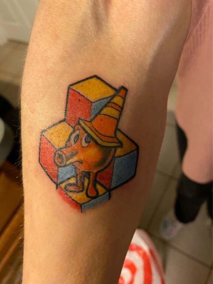 Colorful tattoo of a cartoon character with a long nose and hat on a background of red, yellow, and blue cubes.
