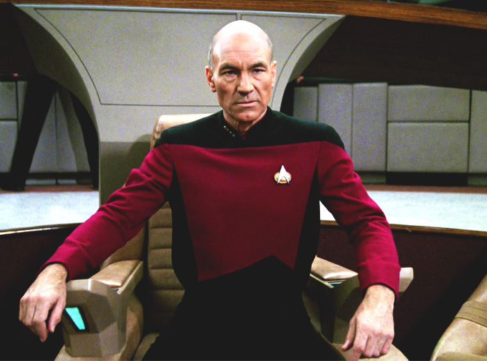 Patrick Stewart in a black and red costume sitting in the armchair