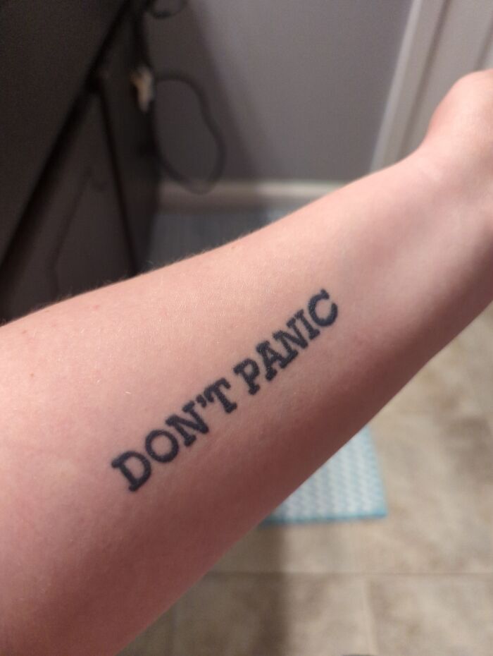 Tattoo of the phrase "DON'T PANIC" on a forearm in black ink.