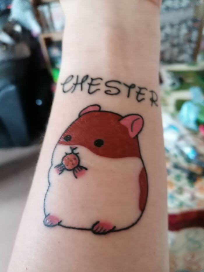 Tattoo of a cute hamster with the name "Chester" on an arm, showcasing tattoos and piercings.