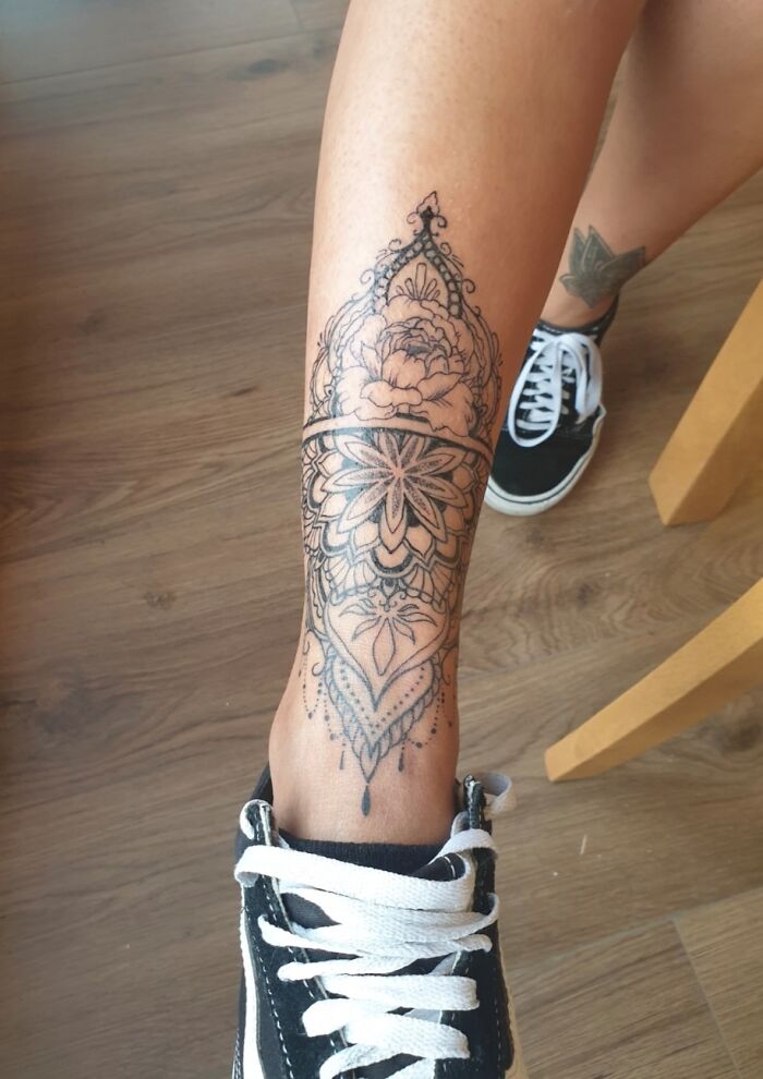 Decorative tattoo featuring floral and geometric elements on a person's leg, with a glimpse of shoes and wooden floor.