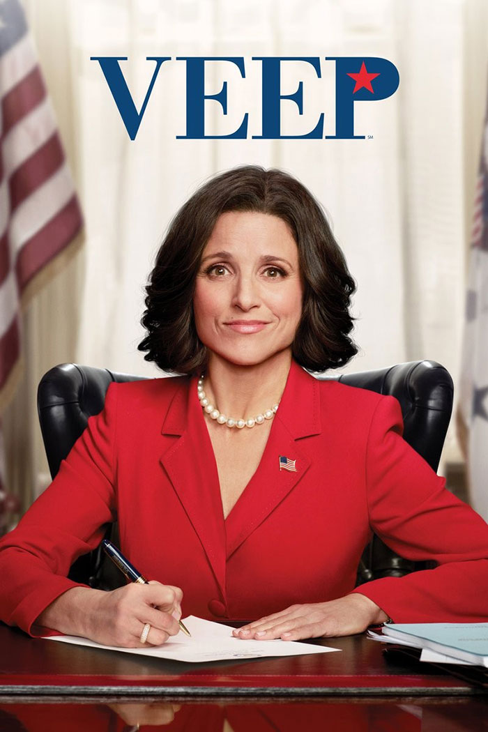 A woman in a red suit writing at a desk, with the "Veep" logo above her head, representing a popular HBO TV show.