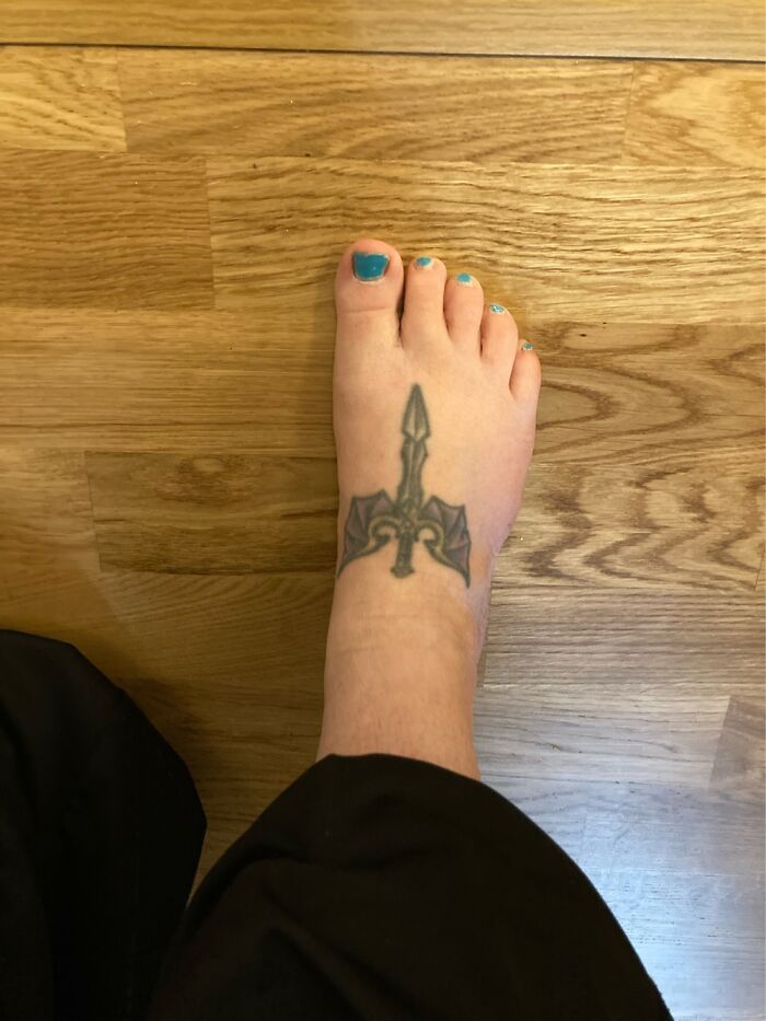 Foot tattoo featuring a sword design, set on wooden flooring.