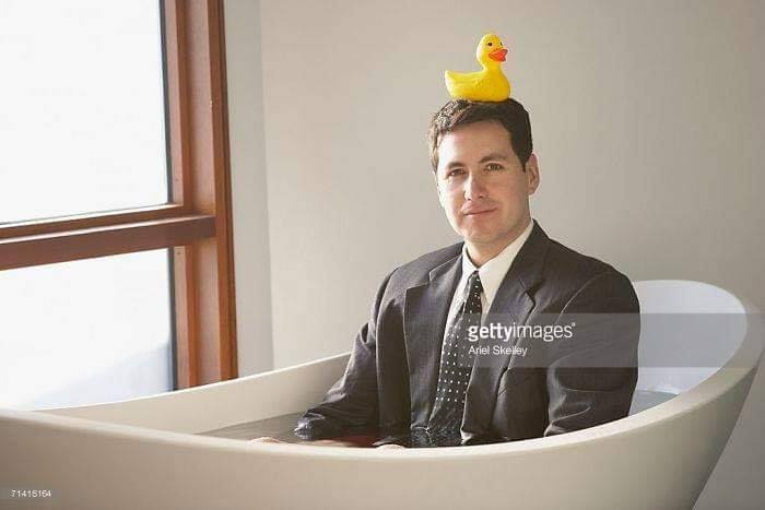 Man in a suit sitting in a bathtub with a rubber duck on his head, capturing weird stock image vibes.