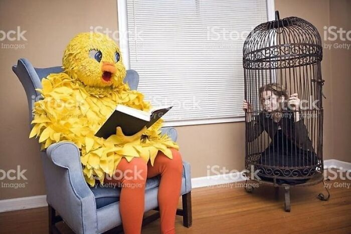 Weird stock image: person in a yellow bird costume reading, while another person is inside a large birdcage in a room.
