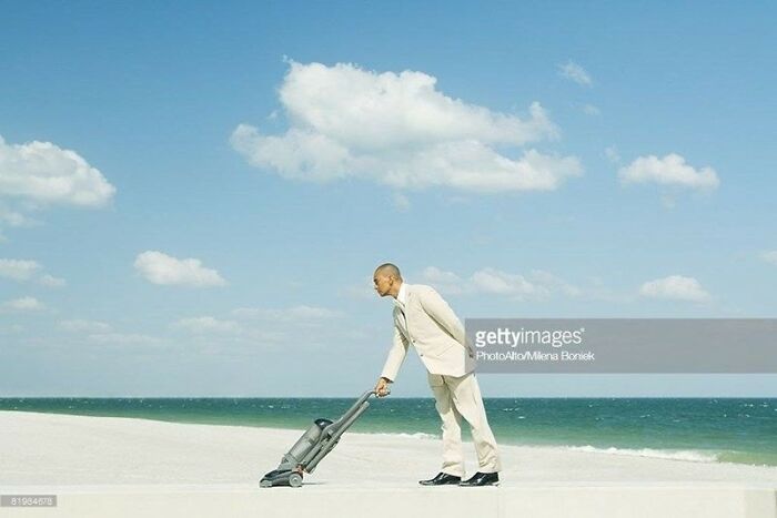 Man vacuuming sand on a beach under a clear blue sky, illustrating a weird stock image concept.