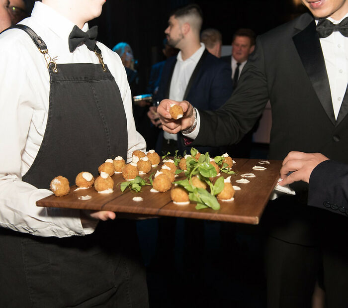 Waiter in black apron serving appetizers to guests in formal attire at an event about big scams shared by an online group