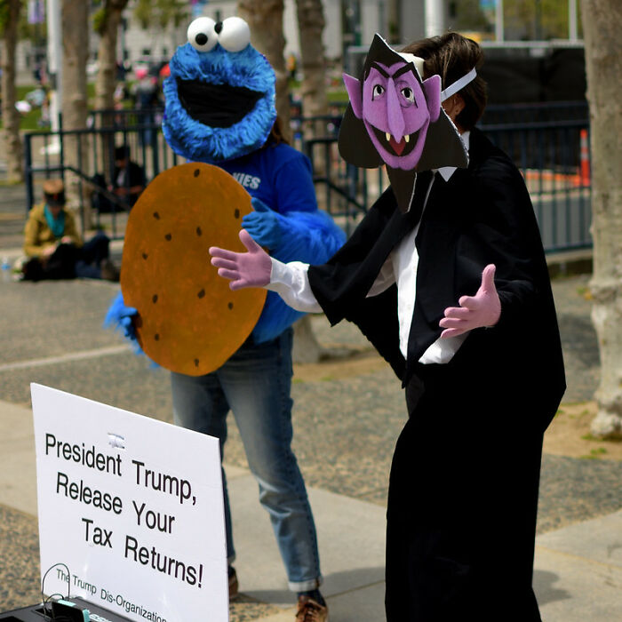People in costumes at a protest highlighting big scams, including a cookie monster and a character with a vampire mask.