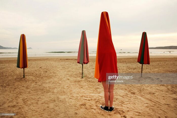 Person standing under an umbrella cover on a beach, surrounded by identical closed umbrellas, depicting a weird stock image.
