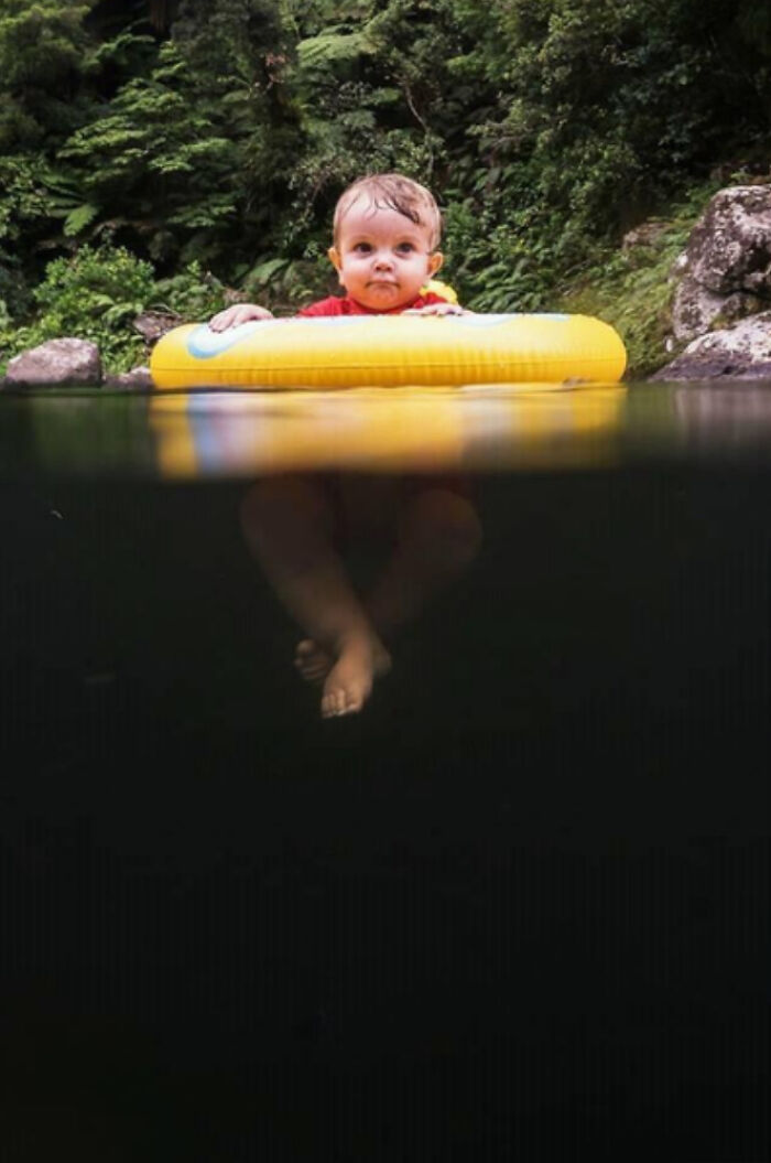 Baby floating on a yellow ring in dark deep water with dense green forest background triggering thalassophobia fears.