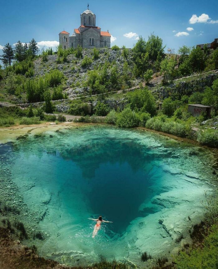 Swimmer near the edge of a deep, clear blue water sinkhole surrounded by rocky hills and greenery triggering thalassophobia fears.