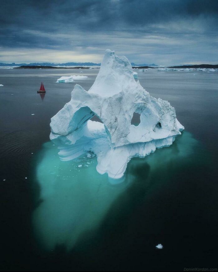 Iceberg floating in deep waters with a small red sailboat nearby, evoking thalassophobia and fear of deep waters.