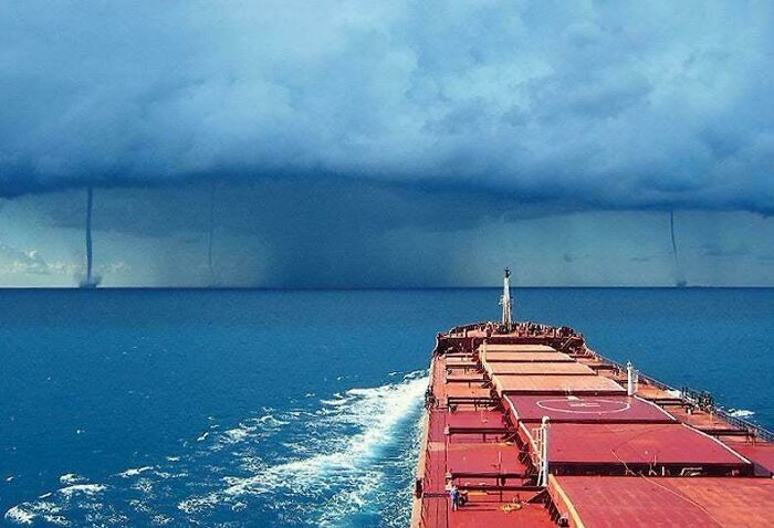 Large cargo ship navigating deep waters under dark storm clouds with multiple waterspouts, triggering thalassophobia fear.