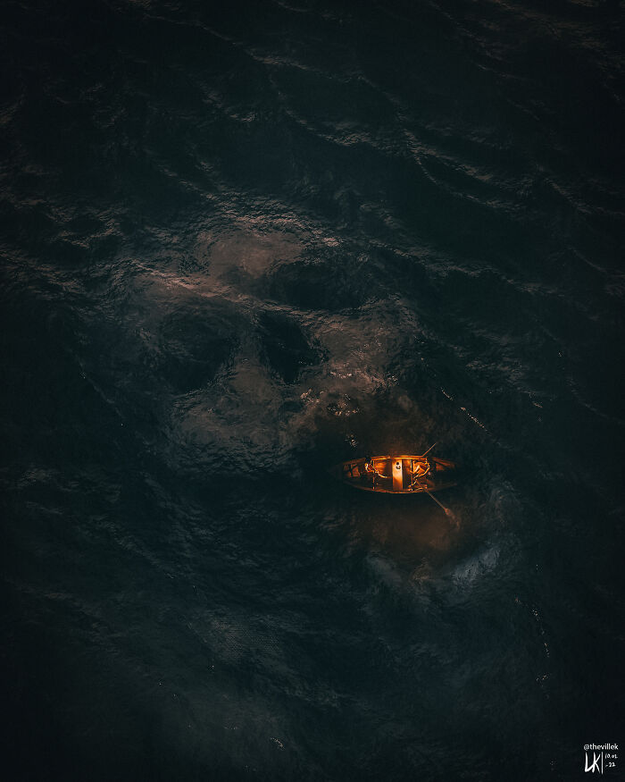 Small illuminated boat floating on dark ocean waters with a subtle skull shape beneath, triggering thalassophobia fears.
