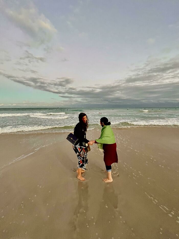 Two women standing barefoot on a sandy beach near the ocean, with waves and cloudy sky triggering thalassophobia fears.