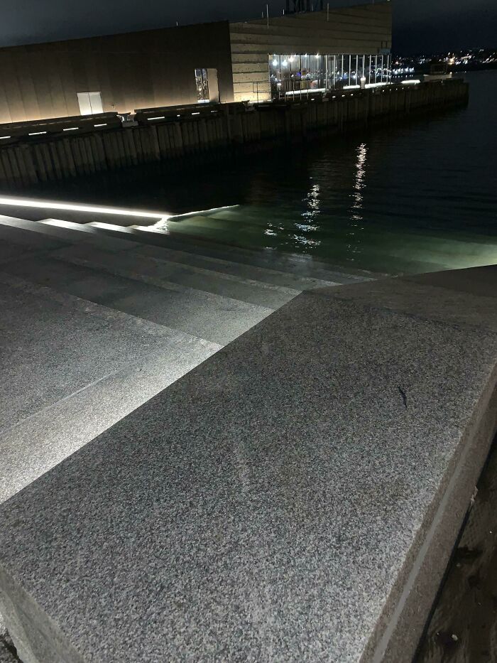 Nighttime view of deep waters near a concrete pier with steps, reflecting lights and evoking thalassophobia feelings.