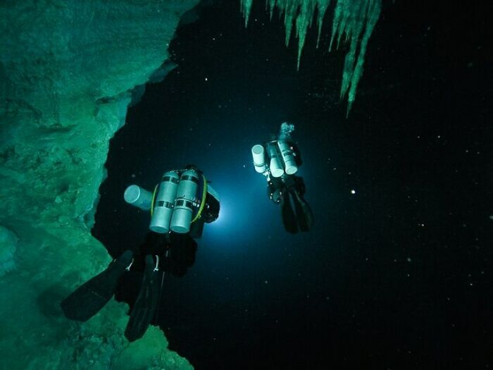 Two scuba divers exploring dark underwater cave with green rock formations, illustrating deep waters thalassophobia trigger.