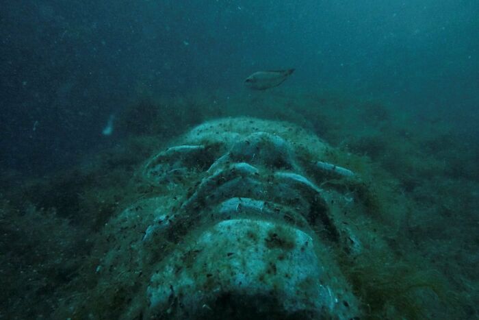 Underwater view of a large ancient statue covered in algae, evoking thalassophobia and fear of deep waters.