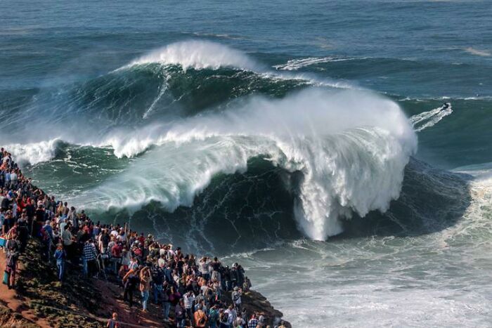 Massive ocean waves crashing near shore with a crowd watching, evoking feelings related to fear of deep waters and thalassophobia.