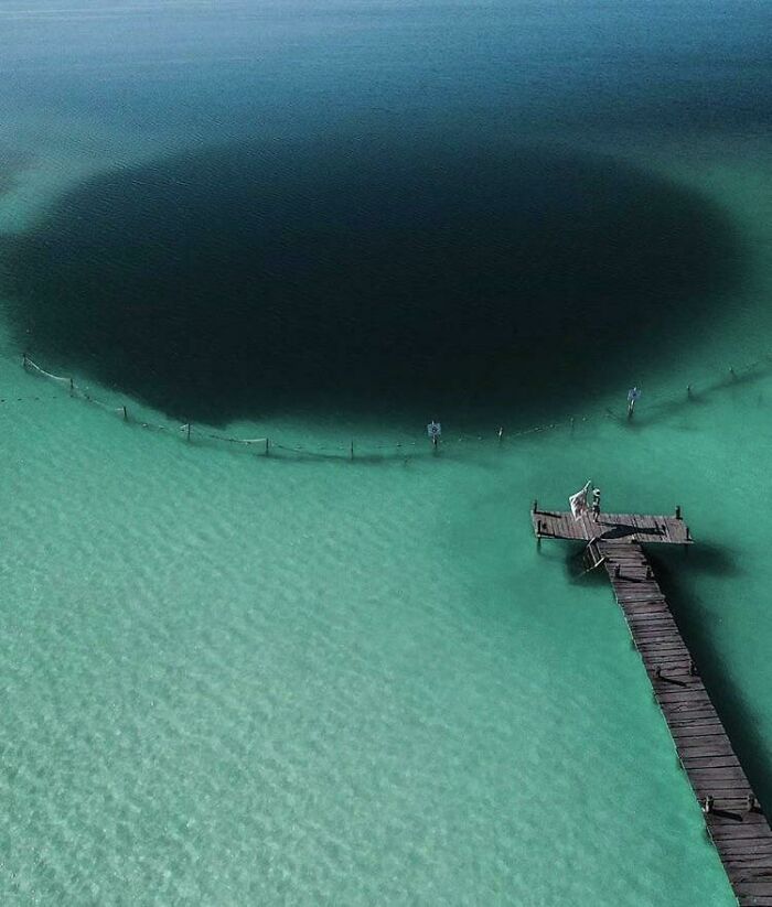 Aerial view of a deep water hole in clear turquoise sea near a wooden dock, triggering thalassophobia feelings.