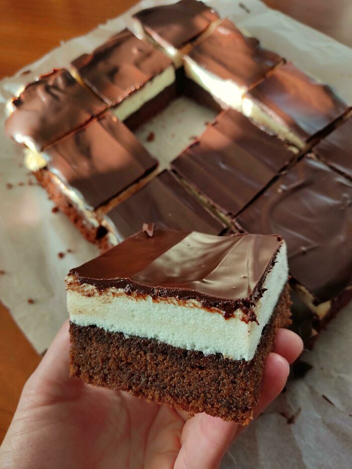 Square piece of nearly perfect layered chocolate dessert with creamy middle and glossy chocolate top held above a tray of similar treats.
