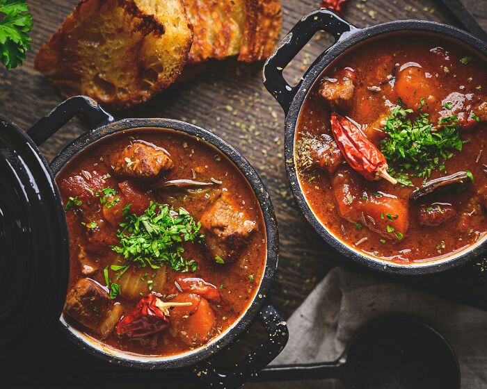 Two bowls of nearly perfect stew garnished with herbs and chili peppers, accompanied by toasted bread slices.