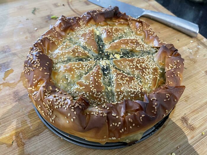 Nearly perfect food photo of a golden baked pie topped with sesame seeds on a wooden surface with a knife nearby.