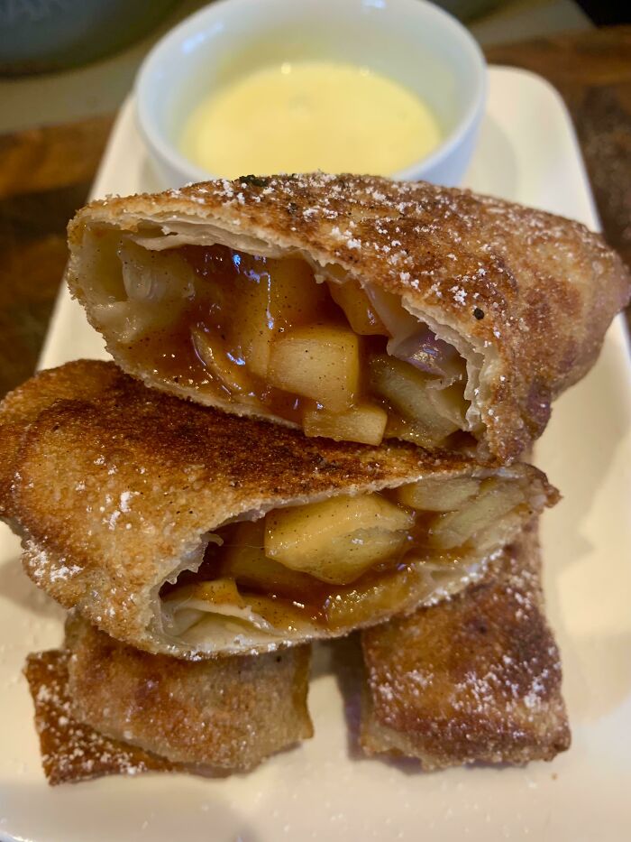 Fried dessert egg rolls filled with warm cinnamon apples, dusted with sugar, served with a creamy dipping sauce, showcasing nearly perfect food.