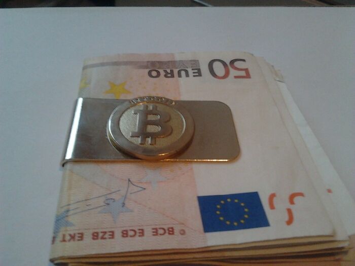 Money clip with Bitcoin symbol on top of folded 50 euro banknotes, illustrating big scams related to cryptocurrency and currency.