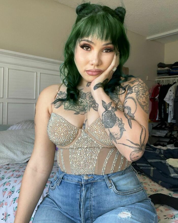 Woman with tattoos and green hair, sitting on bed, looking concerned.
