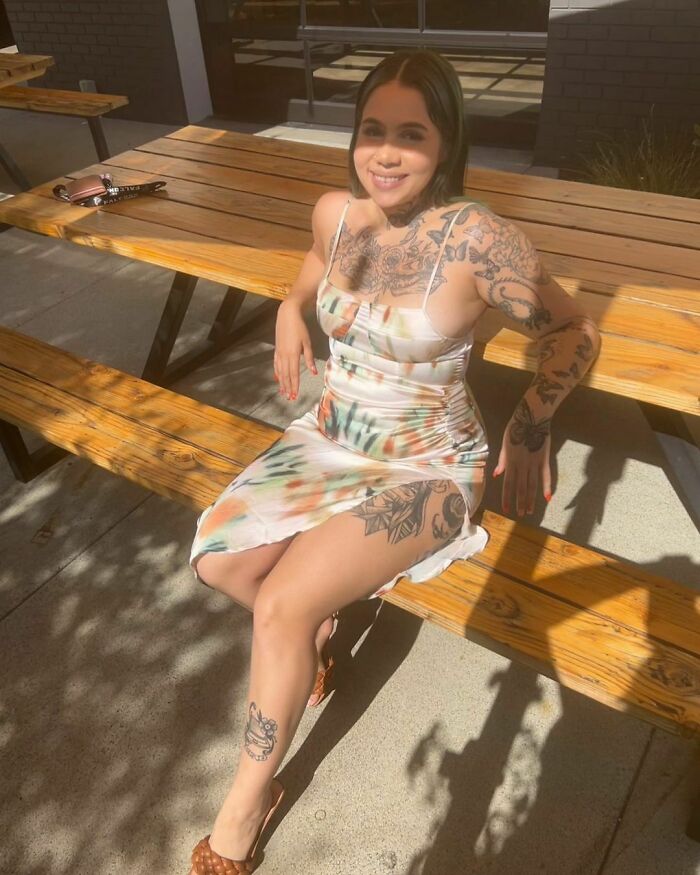 Woman with tattoos sitting on a bench in sunlight, wearing a colorful dress.