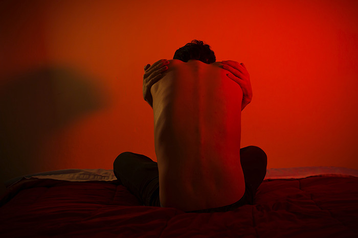 Person sitting on a bed hugging their back under red light, illustrating disturbing things about human bodies concept.