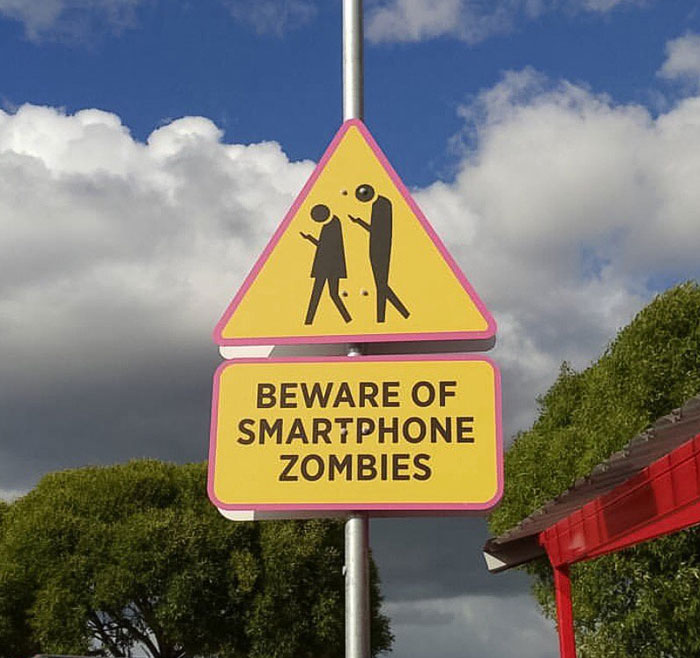 Hilarious and absurd sign warning pedestrians to beware of smartphone zombies on a bright day outdoors.