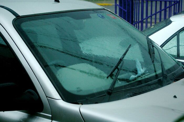 Silver car with a wet windshield and windshield wipers in an outdoor parking lot on a rainy day.
