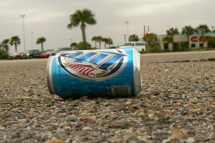 Crushed Miller Lite can lying on rough pavement in a parking lot with palm trees and cars in the background, windshield notes concept.