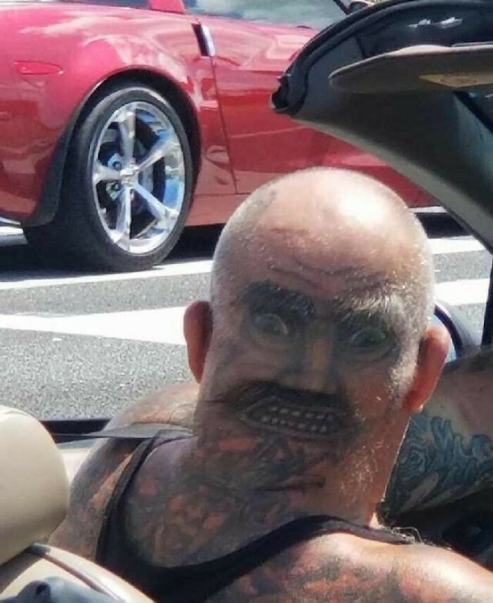 Bizarre tattoo of a face on a bald head, seen in a convertible car.