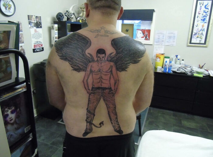 Man with a large, poorly-done tattoo of an angelic figure on his back, featuring dark wings in a tattoo studio.
