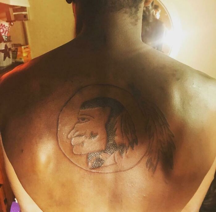 Back tattoo of a poorly-drawn profile portrait inside a circle with feathers.