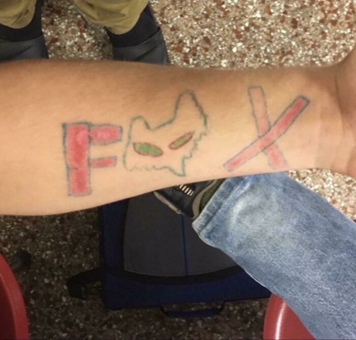 Poorly done tattoo of a fox face with letters "F" and "X" in red and blue ink on a person's forearm.