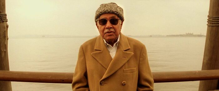 Elderly man wearing sunglasses and a brown coat standing by a wooden railing with a hazy waterfront background, showcasing Easter eggs.