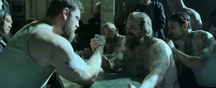 Two muscular tattooed men arm wrestling in a dimly lit room with spectators watching closely, highlighting Easter eggs.