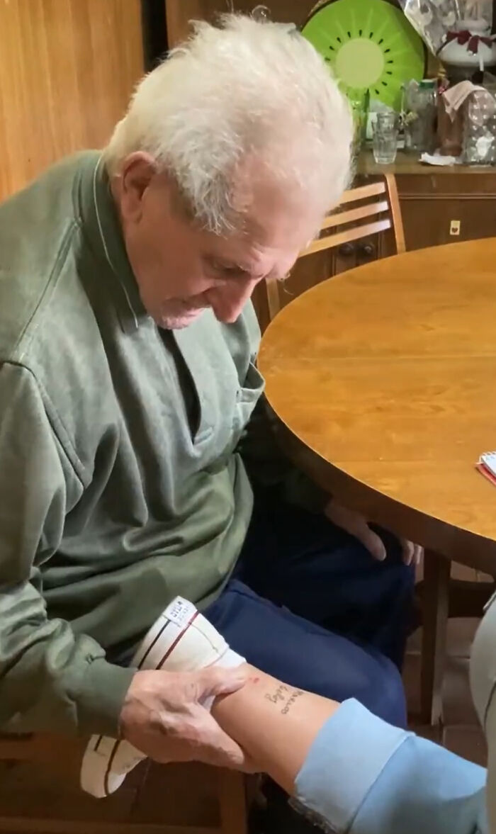 Grandfather reacts emotionally to granddaughter's tattoo, holding her leg at a wooden table.
