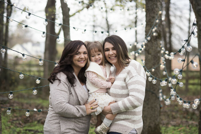A happy family in a forest with string lights, featuring two women and a child, representing LGBTQ+ pride.