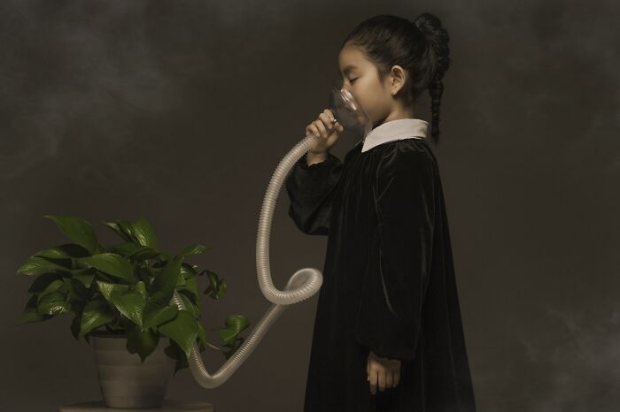 Young girl in a dark dress using a plastic breathing mask connected to a plant, powerful photography concept with smoke.