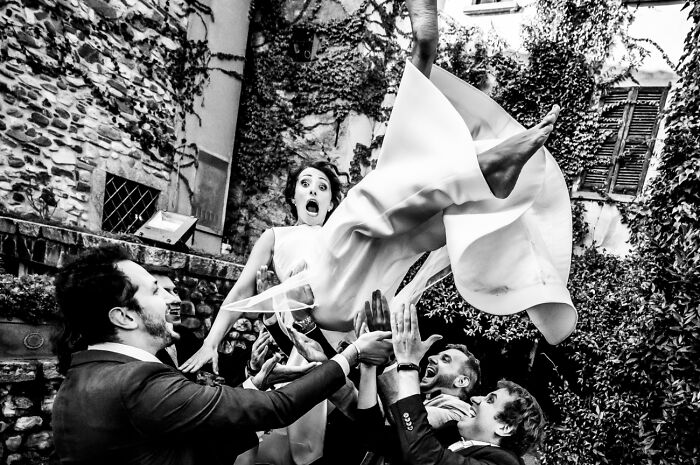 Black and white powerful photography capturing joyful people tossing a woman into the air during a lively celebration outdoors.