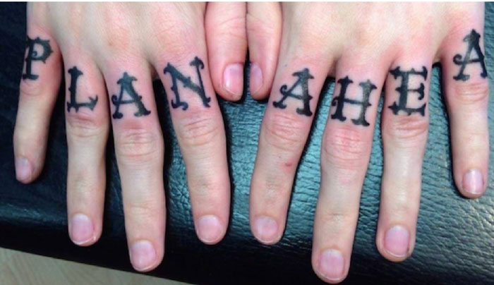 Close-up of hands with misspelled "PLAN AHEA" tattoo on fingers, showcasing tattoo fails.